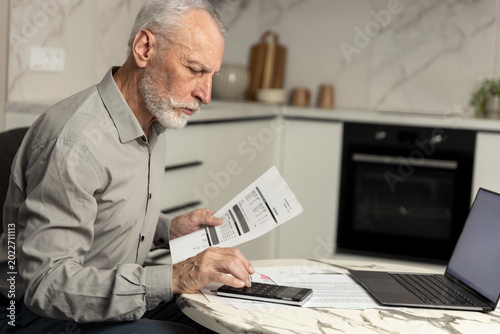 Senior man reviewing utility bills and budgeting at home using his laptop