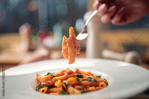 Golden penne pasta glistens with a rich tomato sauce, adorned with vibrant herbs and served on a pristine white plate. A hand lifts a forkful, showcasing the care in every bite