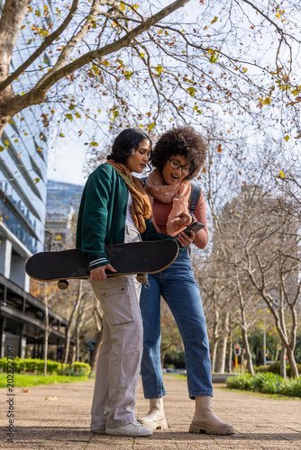 Diverse female friends standing on city path holding skateboard checking smartphone with backpack