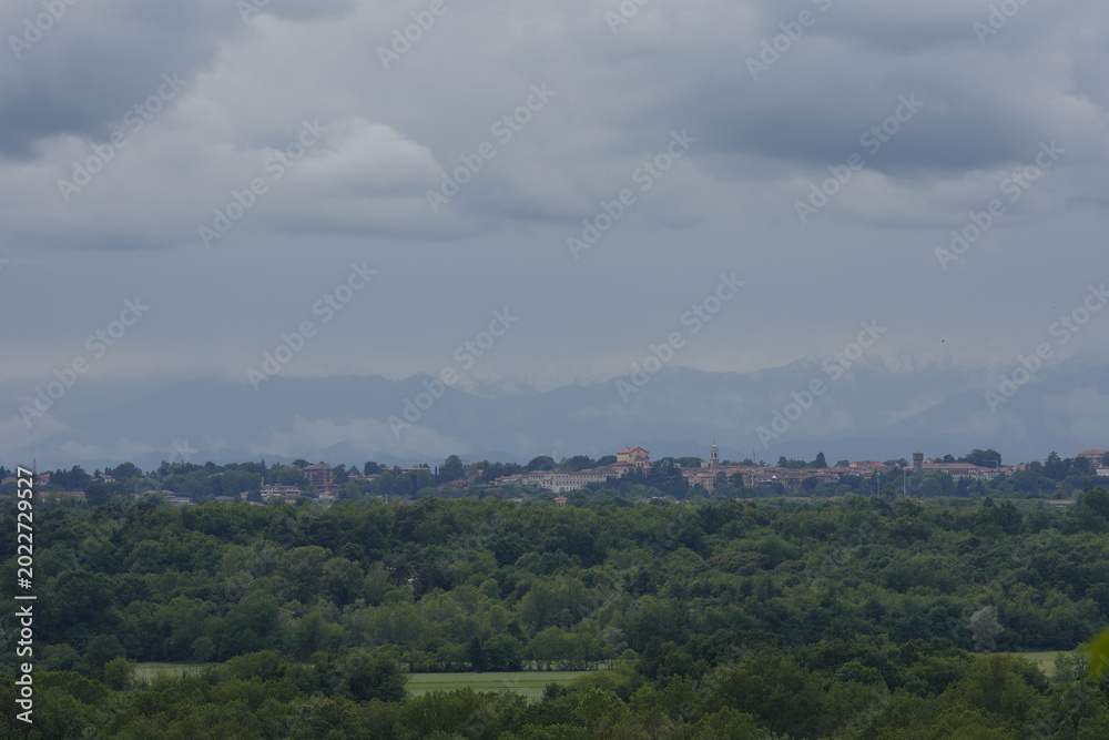 Fototapeta premium A wide landscape shot showing the town of Oleggio visible on the horizon, nestled behind a vast expanse of lush green forest and valley under a bright, cloudy summer sky.