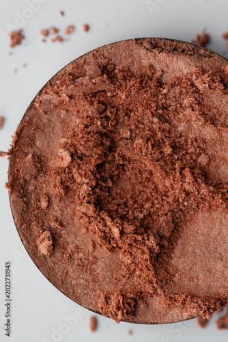 Macro close-up of crushed warm copper bronze eyeshadow powder in a circular makeup pan on white background, shimmering metallic cosmetic pigment with textured beauty powder and scattered makeup