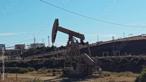 Oil Pump Jack Working In Dry Industrial Field, documentary energy video frame with pipelines, tanks and blue sky backdrop