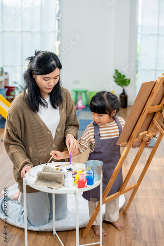 Foto Young asian mom guides preschool daughter selecting paint and brush from supply table near easel