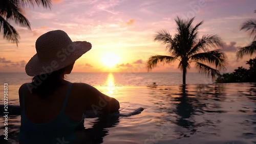 Woman in infinity pool watching tropical sunset behind palm trees