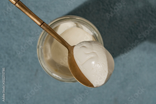 Top view of thick white yogurt or cream cheese or sour cream in glass jar with a golden spoon on a blue background. Minimalist healthy breakfast concept with natural sunlight and shadows
