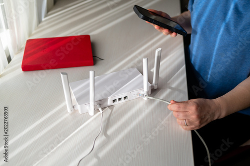Woman Using Smartphone to Configure White WiFi Router While Connecting Ethernet Cable at Home