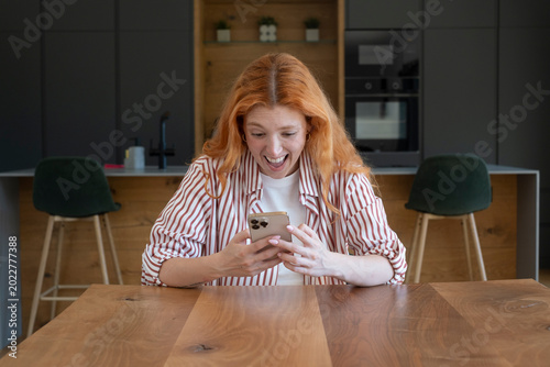 Tableau sur toile Excited redheaded woman reacts to a message on her smartphone while sitting at t