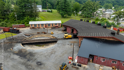 Workers manage train cars and maintenance activities at a roundhouse. The scene features a circular track and various tools around. Green trees and buildings are visible in the background.