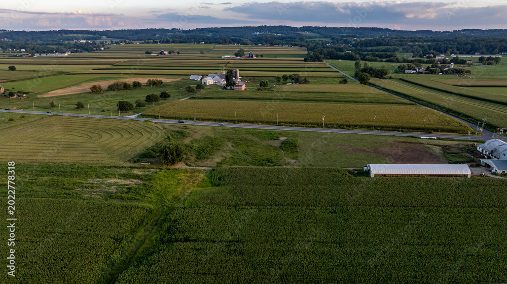 Naklejka premium A vast area of farmland shows green fields stretching out under a colorful sky at sunset. A road runs through the landscape with buildings scattered nearby, highlighting rural life.