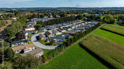 A view shows a cluster of mobile, manufactured homes aligned in rows arranged along a curved road. Nearby, fields stretch out with crops. The scene captures a rural setting in clear weather.