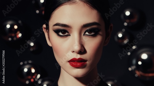 Young asian woman with bold makeup, red lips, dark eyeliner. She looks directly at camera. Background has bokeh lights. Elegant, glamorous, mysterious mood.