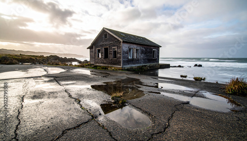 Saltwater pools collect on a cracked asphalt driveway where a coastal bungalow sits abandoned during a high tide event.