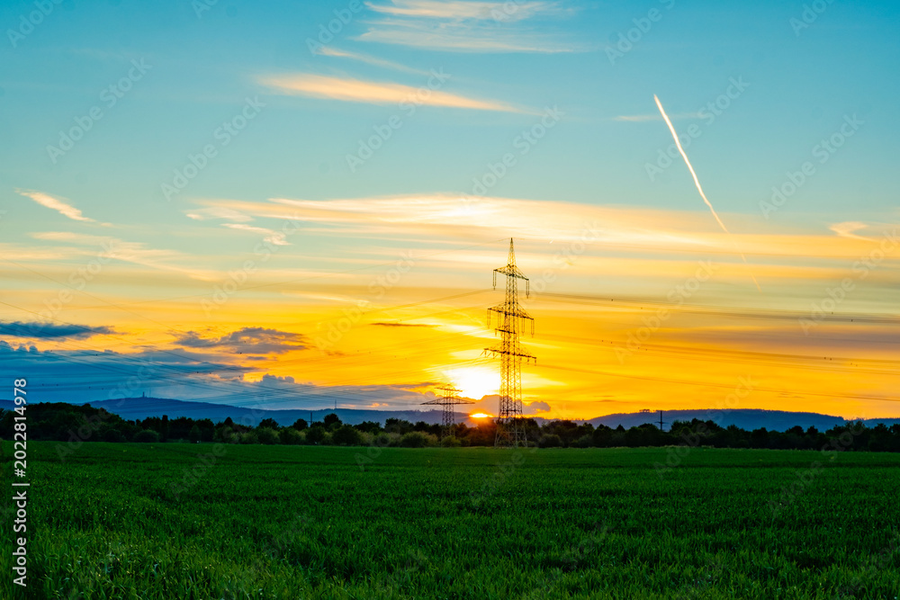 Obraz premium High-voltage transmission towers silhouetted against sunset sky, power lines crossing rural field, Sustainable energy infrastructure, green crops, distant hills, industrial infrastructure meets nature