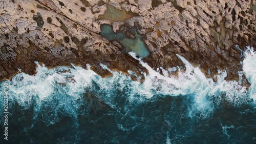 Waves crashing onto a textured rocky shoreline with tide pools from above