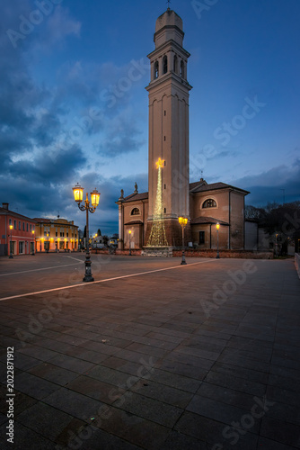 Historic church and bell tower illuminated for Christmas at twilight in the main square of Casier, Treviso, Italy. Beautiful dusk sky over an empty paved piazza 
