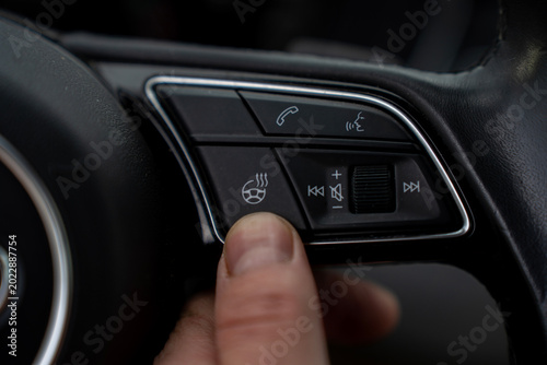 Photos Close up of a finger pressing the steering wheel heater button on a modern car multi-function steering wheel, activating winter comfort features alongside media and phone controls