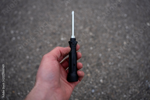 Konstfotografi Hand holding a phillips head screwdriver with a black handle against a blurred asphalt background, representing manual work, repair, or automotive maintenance tools