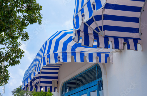 Blue and white striped awnings shade white building with blue window frames under partly cloudy sky and green tree foliage