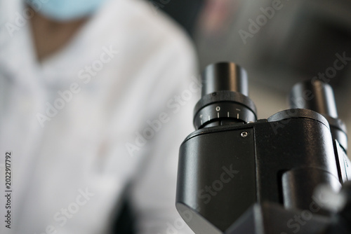 Fotografie Close-up detail of a modern microscope eyepiece with a blurred scientist in a lab coat and mask working in the background