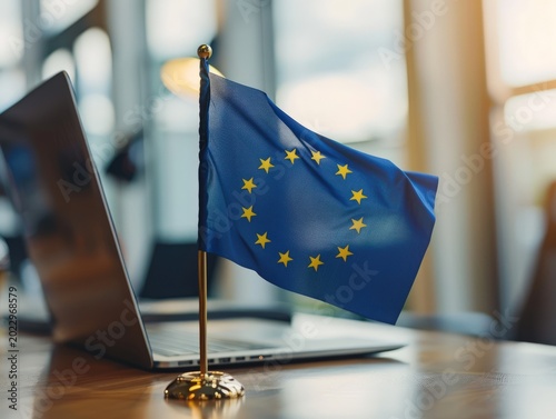 Small blue flag with gold stars standing on a wooden desk next to an open laptop