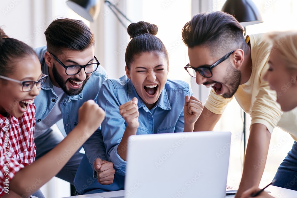 Fototapeta premium Excited young coworkers celebrating success around laptop
