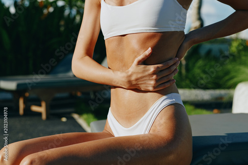 Fotografi A young woman in a white bikini sits by a pool, cupping her toned midriff as sun