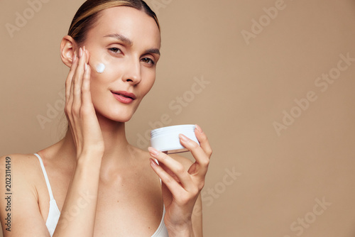 Foto Young woman applies cream to her face, showcasing a simple skincare routine with a smooth glow, neutral background, soft lighting, clear skin, and a calm, focused expression