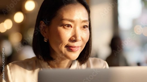 Close up Portrait of an Asian Woman in a White Blouse Working on a Laptop in a Cozy Cafe with Warm Lighting