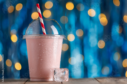 Strawberry milkshake in disposable plastic cup with condensation and red and white striped straw on wooden surface, vibrant bokeh background, refreshing summer treat