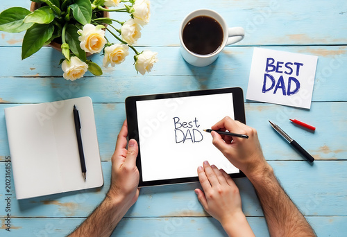 Hands of father and child drawing best dad message on digital tablet on blue wooden table.