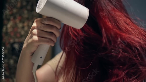 Closeup slow motion of lady in bathroom drying her long red hair with electric appliance. Attractive young adult woman in T-shirt is engaged in her morning routine standing in front of the mirror.