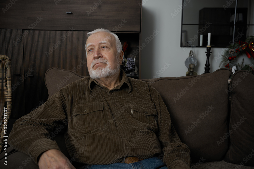 Fototapeta premium Old man sitting on couch in living room during afternoon with relaxed expression and thoughtful gaze