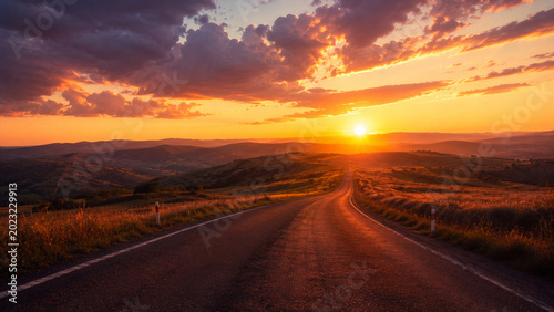 A car journeys along an empty rural highway asphalt road toward the horizon under a beautiful summer sunset sky with clouds over the distant mountain landscape