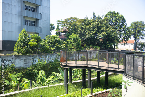 Modern elevated pedestrian walkway in a public park with black steel railings and wooden decking surrounded by lush green trees and a contemporary building facade
