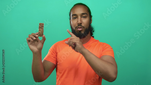 Bild auf Leinwand Man in bright orange shirt holding granola bar and points finger to snack in green studio; health focus