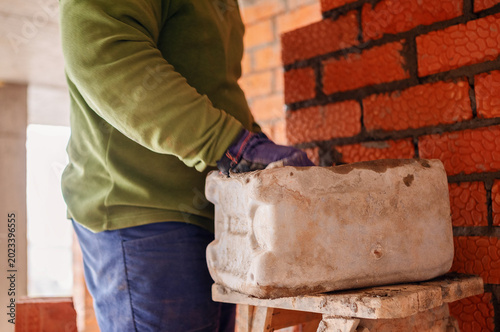 фотографія A construction worker wearing protective gloves reaches into a makeshift mortar tub cut from a plastic jerrycan resting on a stool next to a red brick wall