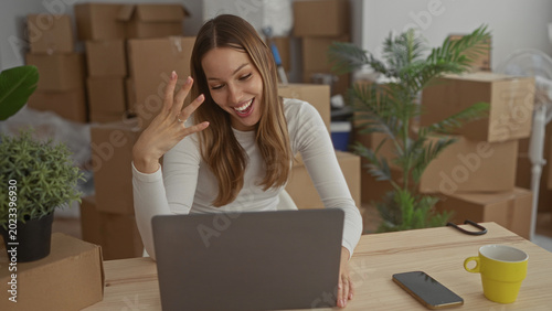 Photos Woman waving hand to laptop in building strewn with moving boxes and houseplants; happiness new beginning
