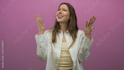 Tableau sur toile Woman young hispanic raising hands and looking upward with pleading expression, wearing white shirt and yellow tanktop in studio with pink wall; frustration yearning