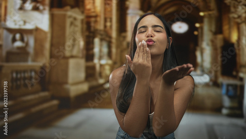 Cuadro en lienzo Young hispanic woman blows a kiss with raised hand to lips in an ornate building hall, puckered lips and casual denim visible; affection warmth