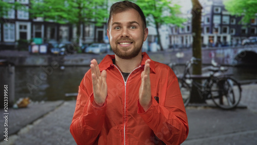 Photography Man in orange prisoner jumpsuit numbered 38258 with palms pressed together praying beside a canal on an amsterdam street near parked bicycles and a bridge; hopeful plea redemption