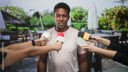 Photos Man speaking into multiple microphones and a smartphone held by reporters on a restaurant patio outside a building; press scrutiny calm