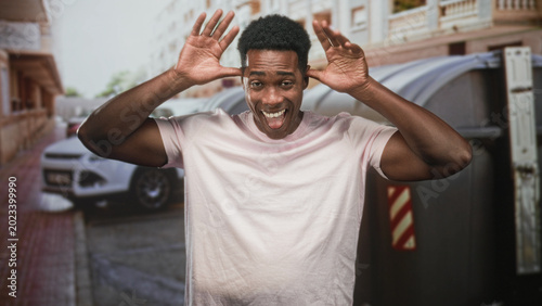 Foto Man raising hands by head and smiling, wearing pink tshirt beside dumpster on street; playful energy mischief