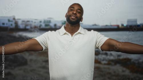 Tableau sur toile Young african american man with hands crossed over chest at seaside building on the beach, bare forearms visible, looking at camera; gratitude welcome