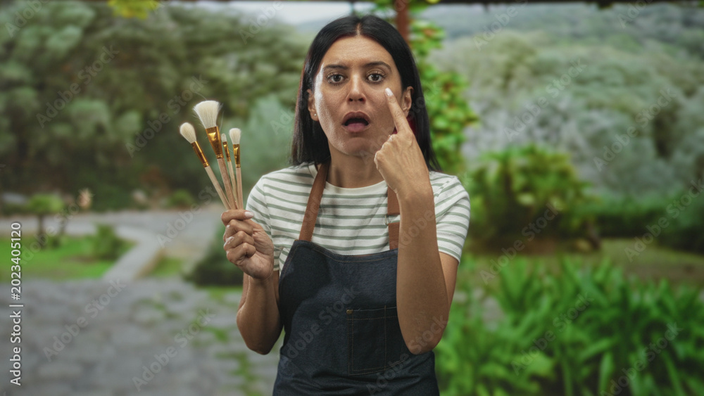 Fototapeta premium Woman wearing apron holding paintbrushes points finger to cheek on a forest path while holding makeup brushes and face cream; serious focus.