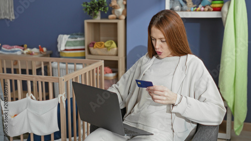 Photos Woman redhead holding a creditcard and using a laptop while seated beside a baby crib and nursery storage shelves in a nursery; parenting multitasking contentment