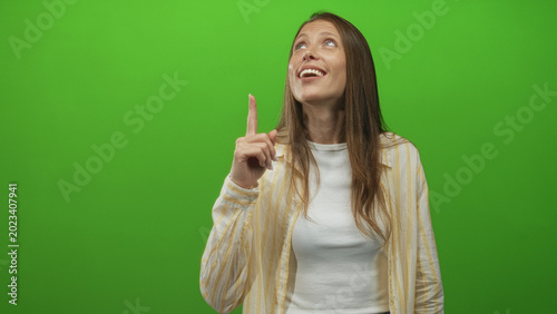 Quadro em tela Young hispanic woman pointing index finger upward in green studio, smiling and looking up while wearing striped shirt; hope