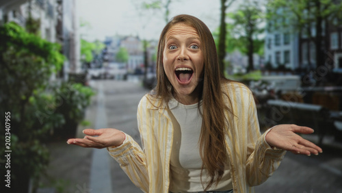 Schilderij op canvas Woman shrugging with open palms on city street near outdoor cafe and bicycles, wearing striped shirt and jeans, expressive face and relaxed posture; lighthearted confusion
