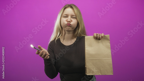 Fotografija Woman holding smartphone and paper bag, puckering lips and holding bag aloft against a purple studio backdrop; playful confidence