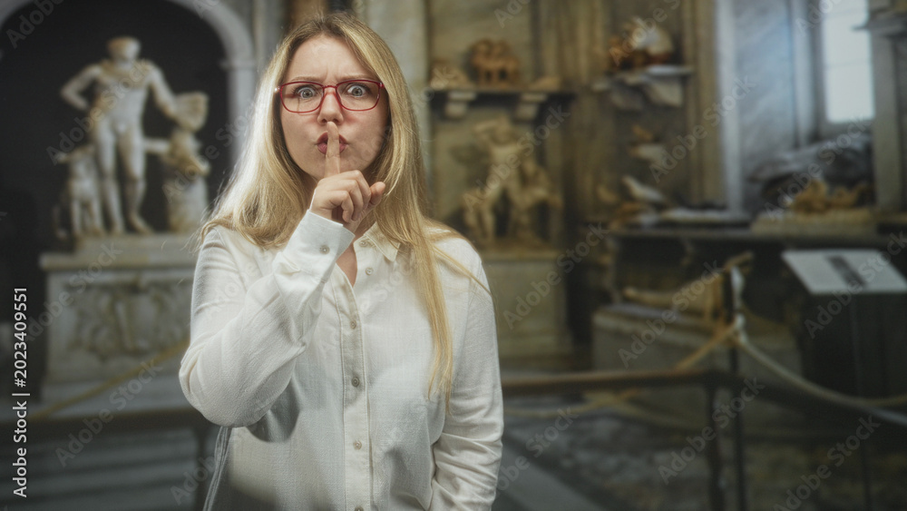 Fototapeta premium Woman wearing red glasses and white shirt with finger to lips for silence and pointing at camera among marble statues and rope barrier in museum; quiet hush respect.