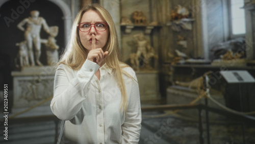 Photos Woman wearing red glasses and white shirt with finger to lips for silence and pointing at camera among marble statues and rope barrier in museum; quiet hush respect